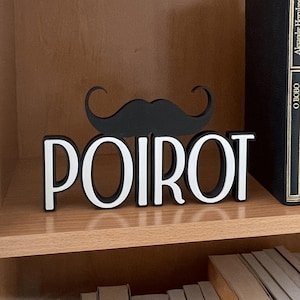 Pu&ograve; includere: Un cartello decorativo in bianco e nero con la scritta "POIROT" e un design a baffi. Il cartello &egrave; su una mensola di legno con libri sullo sfondo.