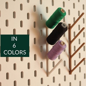 May include: A pegboard with a brown wooden branch-shaped hook holding three spools of thread in green, black, and purple. The text "IN 6 COLORS" is on a green square.