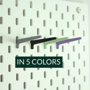 Könnte beinhalten: Fünf Haken in verschiedenen Farben, grau, schwarz, grün und lila, montiert auf einer weißen Lochwand.  IN 5 COLORS steht in schwarzer Schrift unter den Haken.