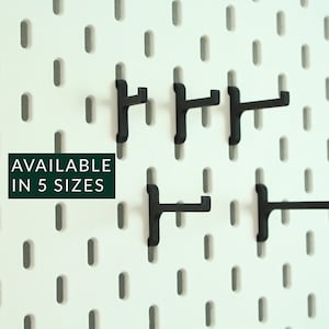 Könnte beinhalten: Fünf schwarze Metallhaken für Lochwand in verschiedenen Größen. Die Haken sind an einer weißen Lochwand befestigt. Der Text "AVAILABLE IN 5 SIZES" ist auf einem grünen Rechteck gedruckt.