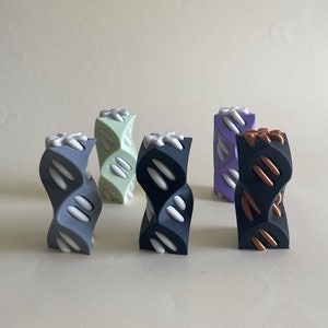 以下が含まれることがあります： 5つの異なる色の3Dプリントされた装飾品。オブジェクトはすべて同じ形、ねじれた円柱で、上部に花のようなデザインが施されています。色はグレー、グリーン、パープル、ブラック、そして銅のアクセントがついたブラックです。