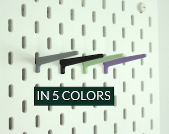 May include: Five peg hooks in different colours, grey, black, green, and purple, mounted on a white pegboard.  IN 5 COLORS is written in black text below the hooks.