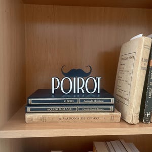 Pu&ograve; includere: Un'esposizione decorativa da scaffale con un baffo nero e il nome "POIROT" in lettere bianche. L'esposizione &egrave; posta sopra una pila di libri con titoli portoghesi, creando un tema letterario.