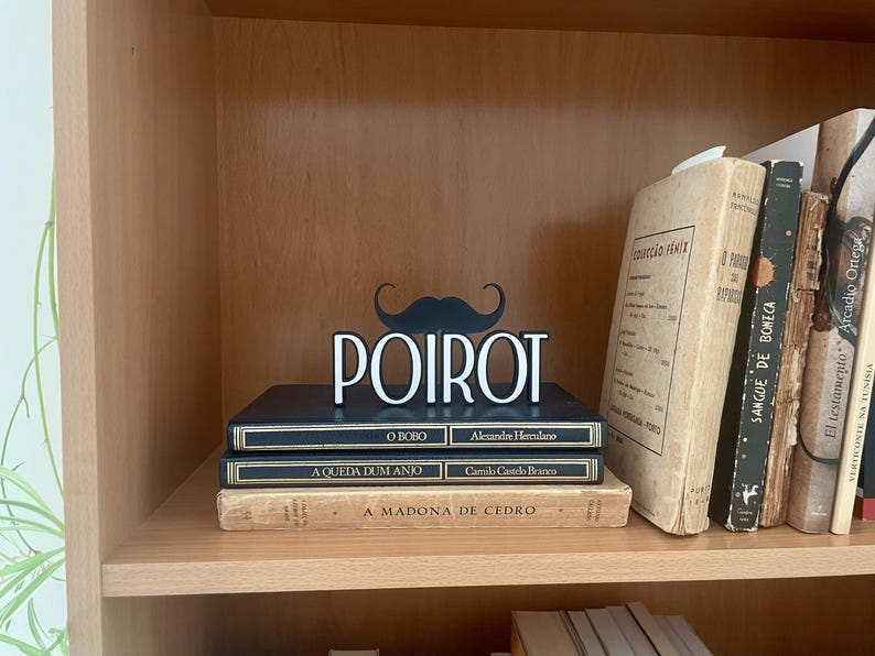 Pu&ograve; includere: Un'esposizione decorativa da scaffale con un baffo nero e il nome "POIROT" in lettere bianche. L'esposizione &egrave; posta sopra una pila di libri con titoli portoghesi, creando un tema letterario.