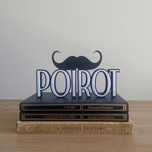 Pu&ograve; includere: Una decorazione a forma di baffi neri e bianchi &egrave; sopra il nome "POIROT" in lettere bianche. Il display poggia su una pila di tre libri con scritte dorate e copertine blu scuro e beige.