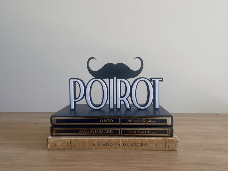 Pu&ograve; includere: Una decorazione a forma di baffi neri e bianchi &egrave; sopra il nome "POIROT" in lettere bianche. Il display poggia su una pila di tre libri con scritte dorate e copertine blu scuro e beige.