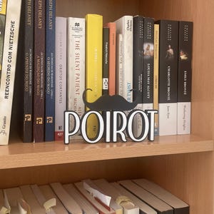 Pu&ograve; includere: Una libreria in legno espone una collezione di libri. Un baffo nero decorativo e la parola "POIROT" in lettere bianche si trovano davanti ai libri. I libri hanno vari colori e titoli.
