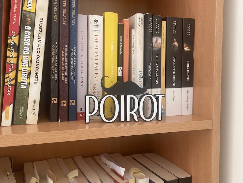Pu&ograve; includere: Una libreria in legno espone una collezione di libri. Un baffo nero decorativo e la parola "POIROT" in lettere bianche si trovano davanti ai libri. I libri hanno vari colori e titoli.
