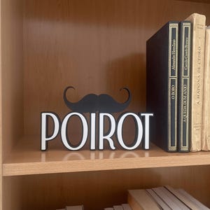 Pu&ograve; includere: Un elemento decorativo da scaffale con il nome "POIROT" in lettere bianche e un baffo nero sopra. Sulla mensola sono visibili libri con scritte dorate, tra cui titoli come "O BOBO" e "A MADONA DE CEDRO".