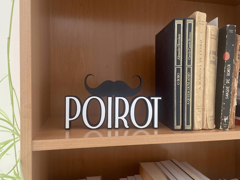 Pu&ograve; includere: Un elemento decorativo da scaffale con il nome "POIROT" in lettere bianche e un baffo nero sopra. Sulla mensola sono visibili libri con scritte dorate, tra cui titoli come "O BOBO" e "A MADONA DE CEDRO".