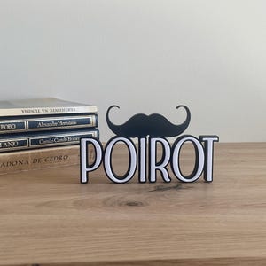 Pu&ograve; includere: Un cartello decorativo in bianco e nero con la scritta "POIROT" e un baffo nero sopra. Il cartello &egrave; su una superficie di legno con libri sullo sfondo.