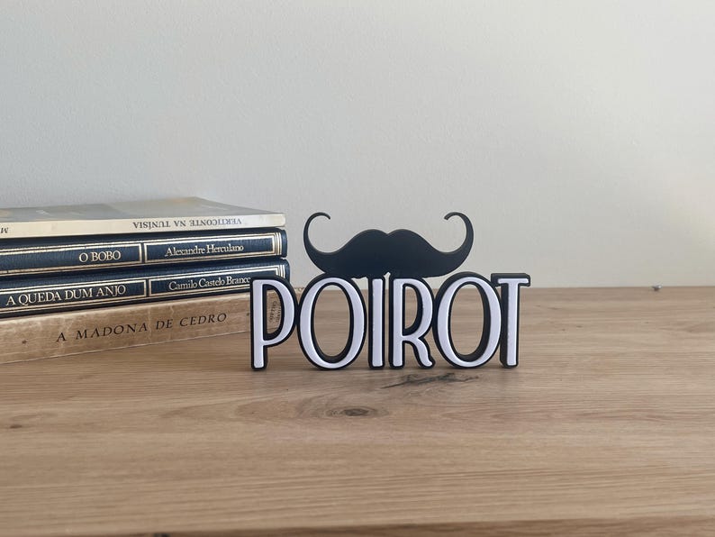 Pu&ograve; includere: Un cartello decorativo in bianco e nero con la scritta "POIROT" e un baffo nero sopra. Il cartello &egrave; su una superficie di legno con libri sullo sfondo.