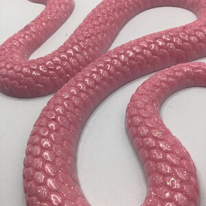 Könnte beinhalten: Eine rosa, glitzerbedeckte Schlangenskulptur mit einem strukturierten, schuppenartigen Muster. Die Schlange ist gewunden und hat einen detaillierten Kopf. Die Figur hat einen leuchtenden Rosaton und eine glänzende Oberfläche.