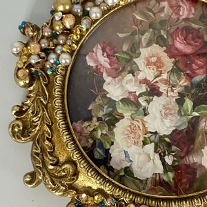 May include: An oval picture frame with a metallic gold finish and ornate scrollwork. The frame is embellished with white pearls, gold beads, and small iridescent crystals. Inside is a print of a bouquet featuring red, pink, and white roses.