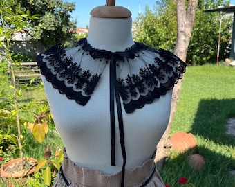 Puede incluir: Un collar de encaje negro con un lazo de cinta negro. El collar está en un maniquí blanco y tiene un borde festoneado.