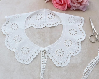 May include: A white, eyelet lace collar with a scalloped edge and floral cutouts. A matching lace trim with small pom-poms is attached. Pink roses and a pair of scissors are in the background. This collar is a delicate accessory.