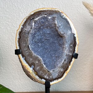 May include: A polished, oval-shaped geode slice, showcasing a deep blue-grey crystal center surrounded by bands of brown and white. The geode is mounted on a black metal stand, perfect for display as a decorative item.