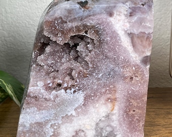 May include: A large, raw amethyst geode with a polished exterior. The crystal features shades of lavender, white, and brown. The geode is displayed on a wooden surface, showcasing its natural beauty. A decorative item for home decor.