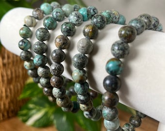 May include: A set of four turquoise gemstone bracelets with dark brown and green speckles. The bracelets are stacked on a white display stand.