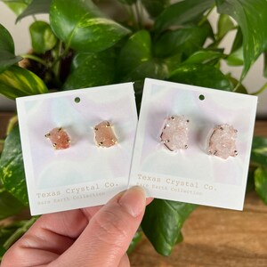 Pu&ograve; includere: Due paia di orecchini di cristallo rosa con dettagli dorati. Gli orecchini sono esposti su cartoncini bianchi con il testo "Texas Crystal Co. Rare Earth Collection".