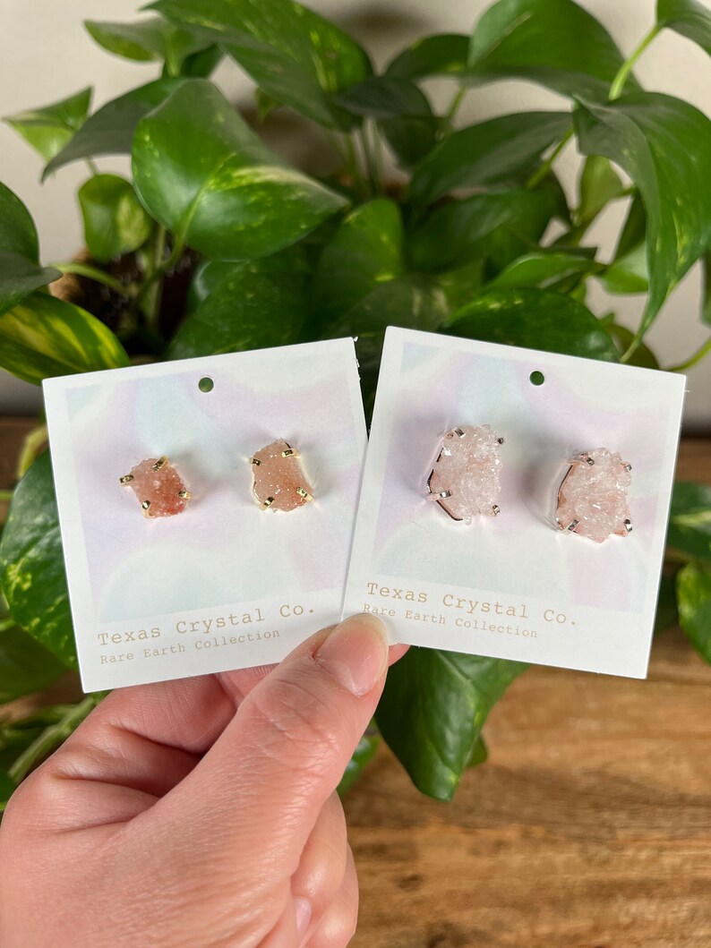 Pu&ograve; includere: Due paia di orecchini di cristallo rosa con dettagli dorati. Gli orecchini sono esposti su cartoncini bianchi con il testo "Texas Crystal Co. Rare Earth Collection".