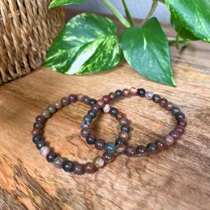 May include: Two bracelets made of brown, red, and green gemstone beads. The bracelets are on a wooden surface.