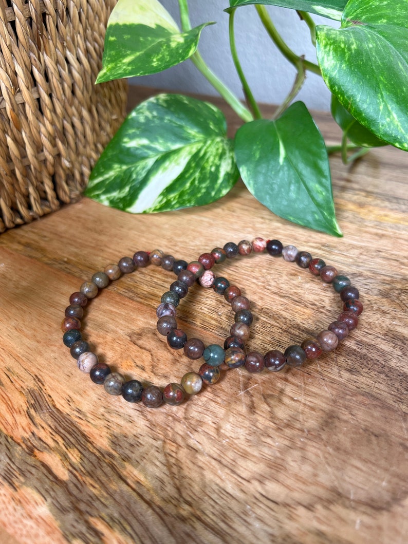 May include: Two bracelets made of brown, red, and green gemstone beads. The bracelets are on a wooden surface.