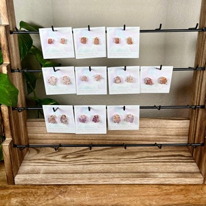 Pu&ograve; includere: Un espositore in legno a tre ripiani presenta una variet&agrave; di orecchini in cristallo rosa e bianco. Ogni paio di orecchini &egrave; esposto su una piccola carta bianca con il testo "Stone Crystal Co."