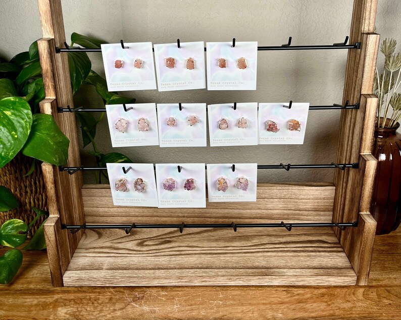 Pu&ograve; includere: Un espositore in legno a tre ripiani presenta una variet&agrave; di orecchini in cristallo rosa e bianco. Ogni paio di orecchini &egrave; esposto su una piccola carta bianca con il testo "Stone Crystal Co."