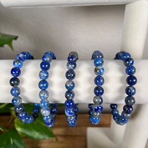 May include: Five blue and grey gemstone bracelets made of round beads. The bracelets are displayed on a white stand.