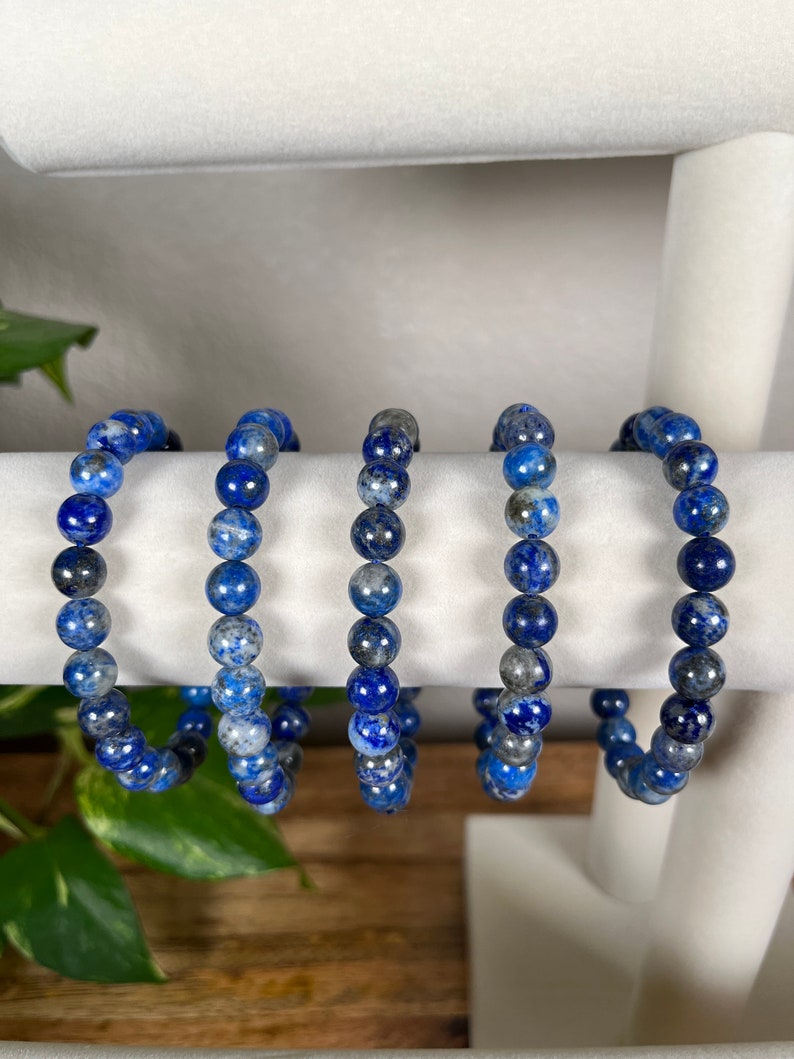 May include: Five blue and grey gemstone bracelets made of round beads. The bracelets are displayed on a white stand.