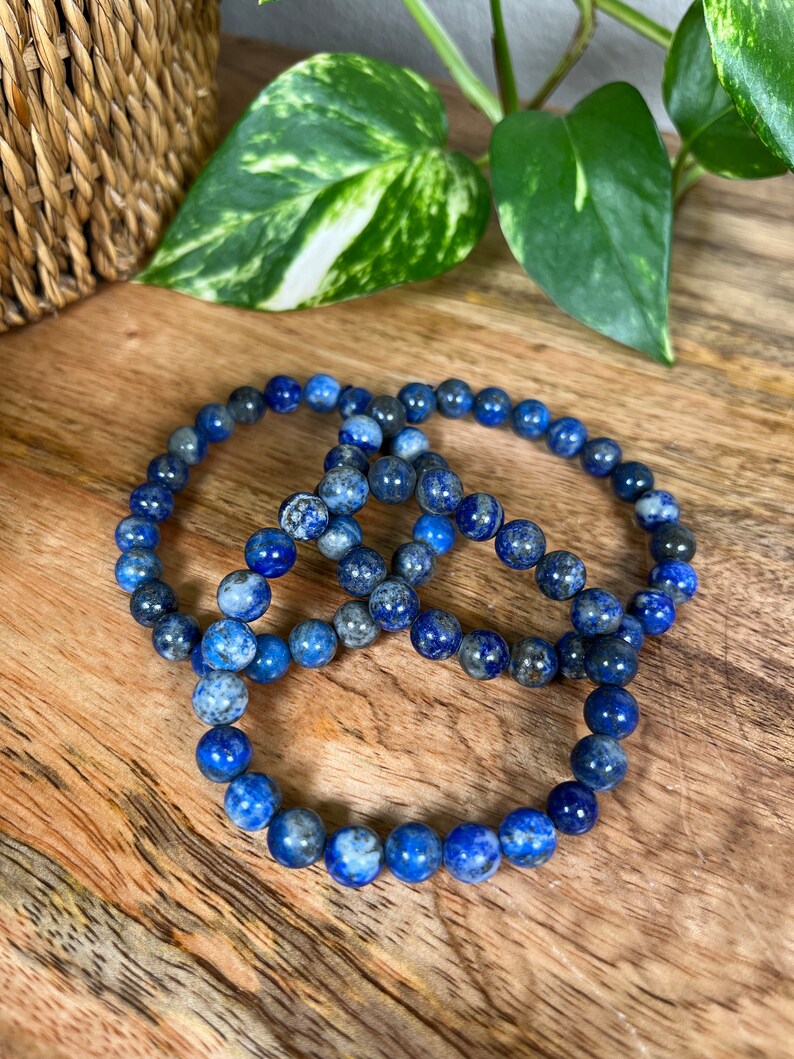 May include: Three blue lapis lazuli beaded bracelets stacked on a wooden surface.
