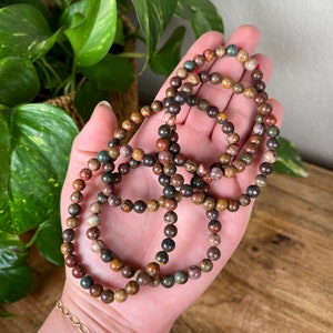 May include: A set of four bracelets made with brown, green, and red jasper beads. The bracelets are stacked on top of each other and are held in a hand.