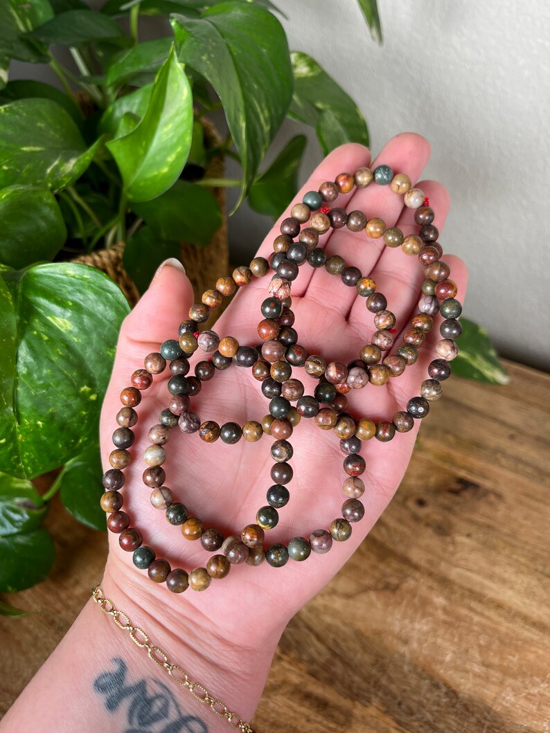 May include: A set of four bracelets made with brown, green, and red jasper beads. The bracelets are stacked on top of each other and are held in a hand.
