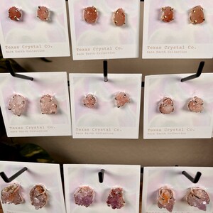 Pu&ograve; includere: Un'esposizione di diversi paia di orecchini con grappoli di cristalli naturali in tonalit&agrave; rosa, arancio e viola. Ogni paio &egrave; montato su un perno dorato ed &egrave; etichettato "Texas Crystal Co. Rare Earth Collection."