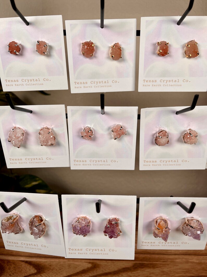 Pu&ograve; includere: Un'esposizione di diversi paia di orecchini con grappoli di cristalli naturali in tonalit&agrave; rosa, arancio e viola. Ogni paio &egrave; montato su un perno dorato ed &egrave; etichettato "Texas Crystal Co. Rare Earth Collection."