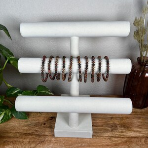 May include: White jewelry display stand with three tiers. The stand is made of wood and has a white fabric covering. There are several brown and red beaded bracelets on the stand.