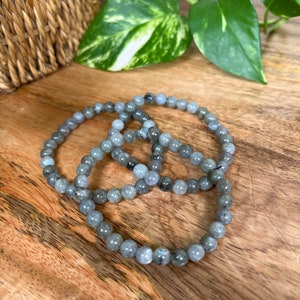May include: Three grey beaded bracelets made of labradorite stones. The bracelets are stacked on top of each other on a wooden surface.