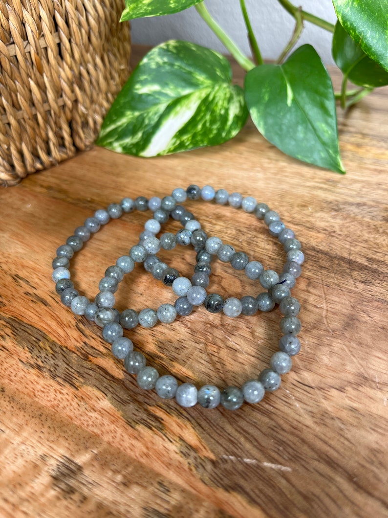 May include: Three grey beaded bracelets made of labradorite stones. The bracelets are stacked on top of each other on a wooden surface.
