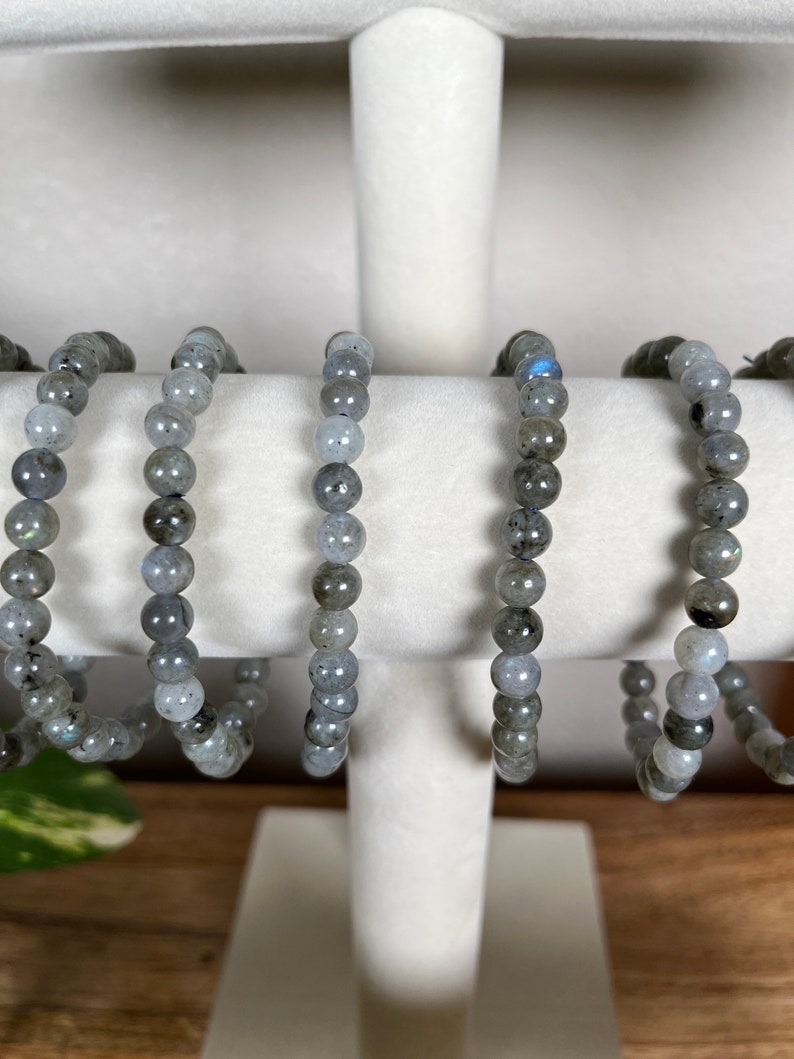 May include: Five grey labradorite beaded bracelets on a white display stand. The bracelets are made of round beads and are all the same size.