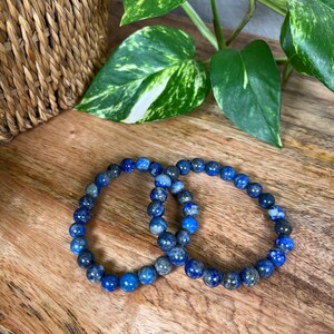 May include: Two blue lapis lazuli beaded bracelets on a wooden surface. The bracelets are made of round beads and are stacked on top of each other.