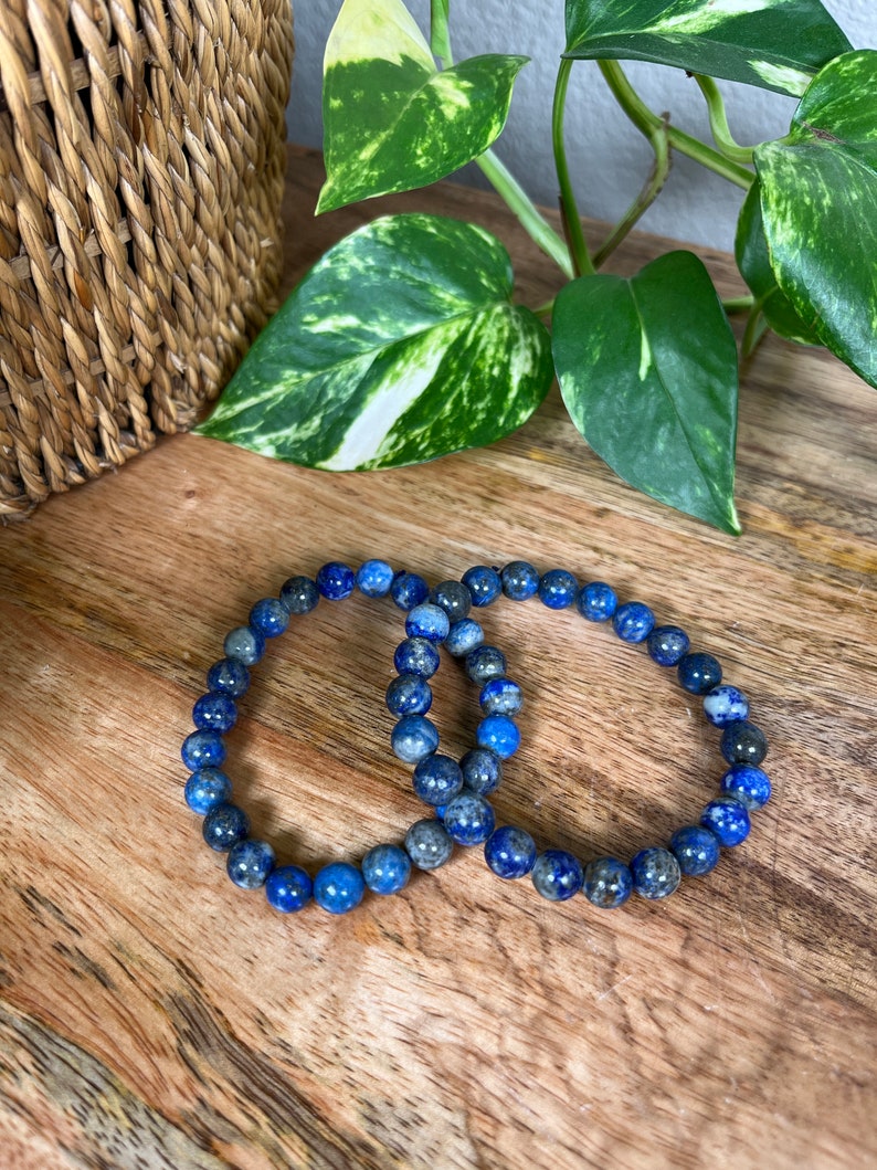 May include: Two blue lapis lazuli beaded bracelets on a wooden surface. The bracelets are made of round beads and are stacked on top of each other.