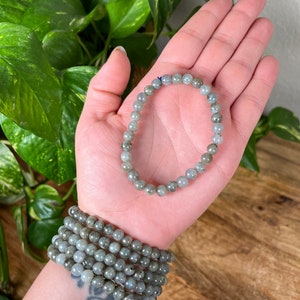 May include: A stack of grey labradorite beaded bracelets. The bracelets are made with round beads and are stacked on a person's wrist.