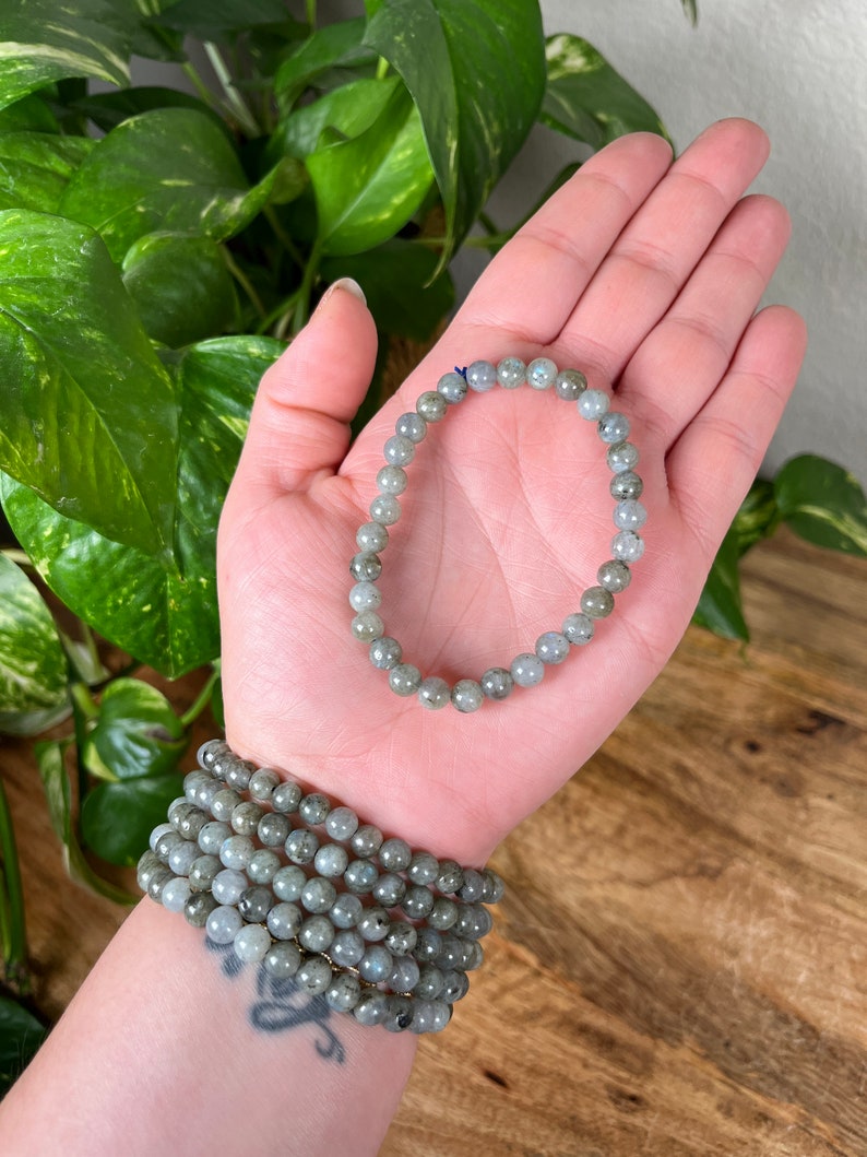 May include: A stack of grey labradorite beaded bracelets. The bracelets are made with round beads and are stacked on a person's wrist.