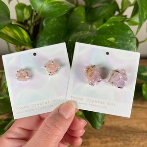 Pu&ograve; includere: Due paia di orecchini di cristallo su carte bianche. Gli orecchini sono realizzati in cristalli rosa e viola e sono incastonati in argento. Le carte riportano il testo "Texas Crystal Co. Rare Earth Collection" stampato su di esse.