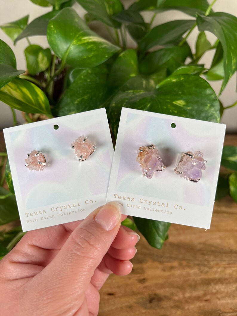Pu&ograve; includere: Due paia di orecchini di cristallo su carte bianche. Gli orecchini sono realizzati in cristalli rosa e viola e sono incastonati in argento. Le carte riportano il testo "Texas Crystal Co. Rare Earth Collection" stampato su di esse.