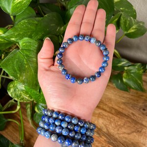 May include: A stack of blue lapis lazuli beaded bracelets. The bracelets are made with round beads and have a dark blue color with flecks of gold.