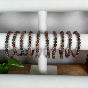 May include: A set of eight bracelets made with brown, red, and gray gemstone beads. The bracelets are displayed on a white jewelry stand.