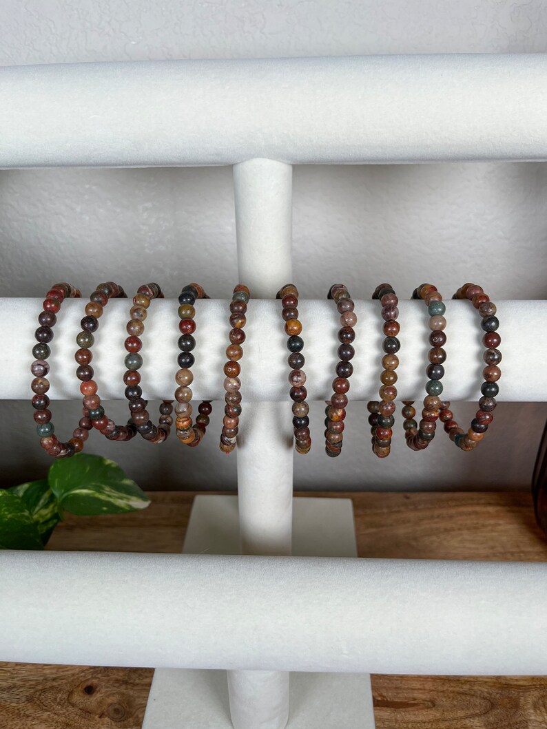 May include: A set of eight bracelets made with brown, red, and gray gemstone beads. The bracelets are displayed on a white jewelry stand.