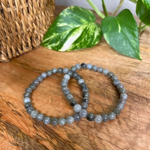 May include: Two grey beaded bracelets made of labradorite stones. The bracelets are stacked on top of each other on a wooden surface.
